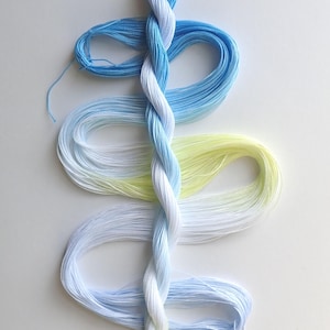 May include: A skein of embroidery floss in shades of blue, white, and yellow, twisted together. The floss is arranged in a spiral pattern, with the colors blending from light to dark. The image is taken from above, showing the full length of the floss.