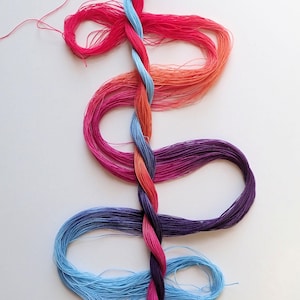 Puede incluir: Una colorida exhibición de hilo de bordar en tonos rojos, naranjas, azules y morados. Los hilos están entrelazados en un patrón en espiral, creando un diseño visualmente atractivo. La imagen está sobre un fondo blanco.