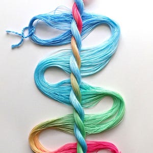 May include: A braided bundle of embroidery floss featuring a gradient of colors including bright pink, light blue, soft yellow, and mint green. The strands are twisted into a central plait with loose, flowing loops extending outward on both sides.