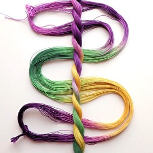 May include: A colorful display of embroidery floss in shades of purple, green, and yellow, arranged in a decorative pattern. The floss is twisted in the center, creating a braided effect. The image is set against a white background.