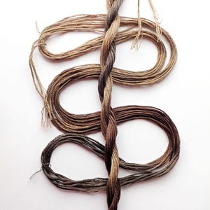 May include: A skein of embroidery floss in various shades of brown, twisted together to form a rope-like structure. The floss is arranged in a looping pattern against a white background, with loose strands extending from the top.