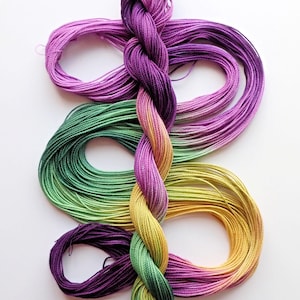 May include: A skein of embroidery floss in a gradient of colors, including purple, green, and yellow. The floss is twisted into a braid, with loose strands forming loops. The image is on a white background.