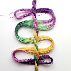 May include: A colorful arrangement of embroidery floss in shades of purple, green, and yellow. The floss is twisted and looped, creating a decorative design. The image is set against a white background.