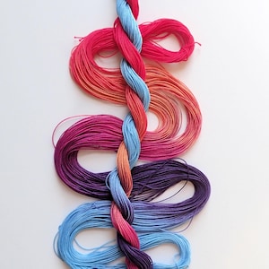 May include: A colorful skein of embroidery floss, featuring a braided section in shades of red, pink, orange, blue, and purple. The floss is arranged in a wavy pattern against a white background. The image includes the text "yarnplayer.etsy.com".