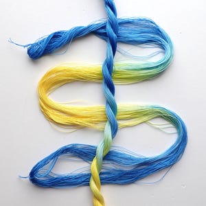 May include: A length of twisted embroidery floss featuring a gradient transition from bright blue to yellow. The floss is arranged in a dollar sign shape, with the vertical bar formed by a tightly twisted strand and the curves by loose loops.