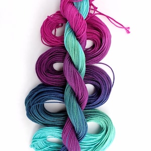May include: A vertical composition of colorful yarn skeins in shades of teal, purple, and blue. The yarn is twisted in the center, creating a gradient effect. The skeins are arranged on a white background, with the Etsy shop name in the bottom right corner.