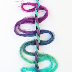 May include: A collection of embroidery floss spools in shades of purple, blue, and green. The floss is arranged in a vertical line, with the darkest color at the top and the lightest color at the bottom. The floss is twisted together in a braid.