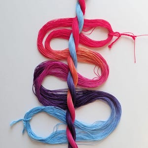 May include: A colorful display of embroidery floss in shades of pink, orange, purple, and blue. The floss is arranged in a vertical pattern, with a twisted central strand and looped sections. The image is set against a white background.