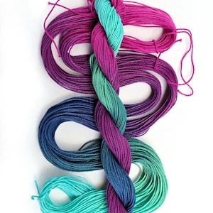 Peut inclure: Un écheveau de fil à broder coloré dans les tons de turquoise, bleu, violet et rose. Le fil est torsadé en tresse, avec des brins lâches qui s'évasent. Le fond est blanc.