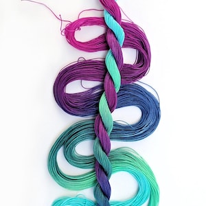 May include: A braided strand of colorful embroidery floss in shades of teal, green, blue, purple, and pink. The floss is arranged in a spiral pattern against a white background, showcasing the vibrant colors.