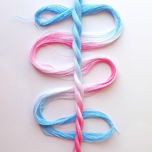May include: A length of embroidery thread featuring a gradient color transition from light blue to white to pink. The thread is twisted into a central vertical braid with loose, curved loops extending outward on both sides of the central strand.