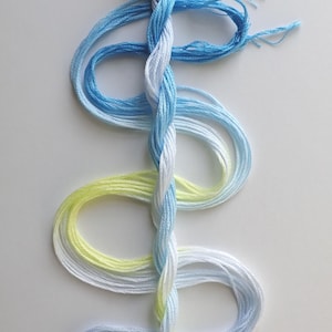 May include: A skein of embroidery floss in shades of blue, white, and yellow. The floss is twisted together, creating a gradient effect. The colors transition from light blue to white, then to yellow. The image is on a white background.