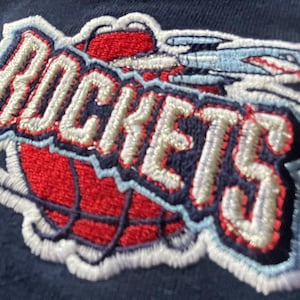 Puede incluir: Parche bordado con la palabra "ROCKETS" en puntadas blancas sobre un fondo azul. El parche presenta una pelota de baloncesto roja con puntadas blancas y una nave espacial azul y blanca.