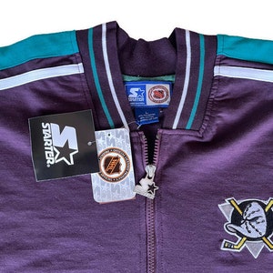 Op de afbeelding: Een paarse Starter-jas met teal en witte accenten. De jas heeft een volledige ritssluiting, een kraag en het Anaheim Ducks-logo op de borst. Het Starter-logo en het NHL-label zijn zichtbaar.
