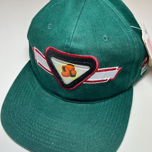 Puede incluir: Una gorra de béisbol verde con un parche blanco y rojo que presenta un personaje de dibujos animados. El parche tiene forma de triángulo con un borde negro.