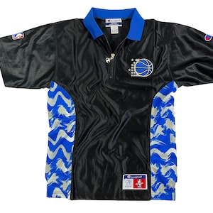 Può includere: Maglia nera a maniche corte con colletto e dettagli blu. Presenta una grafica di basket con stelle e chiusura con zip. La maglia ha motivi a pennellate blu e bianche sui lati e un logo Champion.