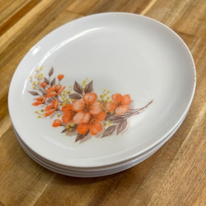 May include: A stack of white ceramic plates with a floral design. The plates feature orange flowers with brown and yellow accents. The plates are stacked on a wooden surface.