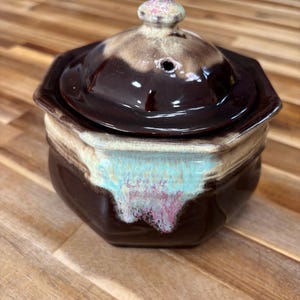 May include: A hexagonal ceramic container with a lid. The container is primarily brown with a turquoise and pink accent. The lid has a small knob and a hole. The container is likely for food storage.