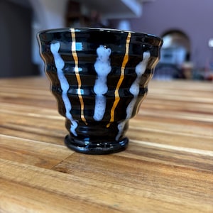 May include: A black ceramic cup with white and gold accents. The cup has a textured, wavy design and is decorated with vertical gold stripes.