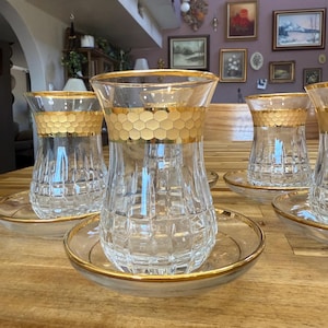 May include: A set of clear glass tea cups and matching saucers, each featuring a gold-rimmed honeycomb pattern around the upper section and a faceted, grid-like texture on the lower body. The glass edges and saucer rims are trimmed in gold.