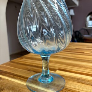 May include: A light blue, swirl-patterned glass brandy snifter. The glass has a rounded bowl and a stem, with a circular base. The glass is sitting on a wooden surface.