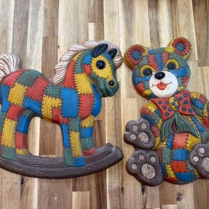 May include: Two wall plaques featuring a rocking horse and a teddy bear, both decorated with a colorful patchwork pattern of red, blue, yellow, and green squares. The rocking horse has a white mane and tail, while the bear wears a red bow tie.