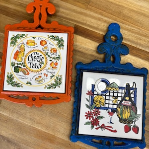 May include: Two decorative trivets. One is orange with a white tile featuring cheese varieties and the text "The Cheese Table." The other is blue with a tile showing cheese, wine, and strawberries. Both have ornate handles.