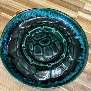 May include: A round, ceramic ashtray with a dark green and teal glaze. The ashtray has a central recessed area with a textured pattern and a surrounding rim. The outer edge has a teal, textured finish.