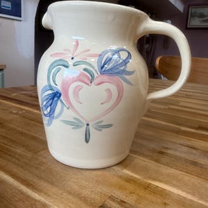May include: A cream-colored ceramic pitcher featuring a hand-painted design of a central pink heart surrounded by blue and green floral accents. The pitcher has a curved handle on the right side and a pouring spout at the top rim of the vessel.