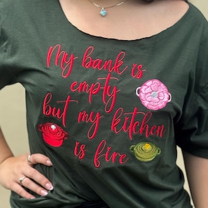 以下が含まれることがあります： 赤い筆記体で「My bank is empty but my kitchen is fire」と刺繍された、濃い緑色のTシャツ。赤、ピンク、緑色の刺繍された鍋のグラフィックが特徴です。