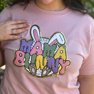 Puede incluir: Camiseta rosa con un parche bordado de colores que dice "Mama Bunny" con orejas de conejo y huellas de patas.