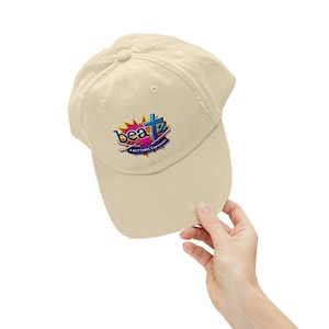 May include: A beige baseball cap with a distressed brim. The cap features a colorful logo that reads "beatz" with a cross design and the text "A RHYTHMIC Expression". The cap is a casual accessory.