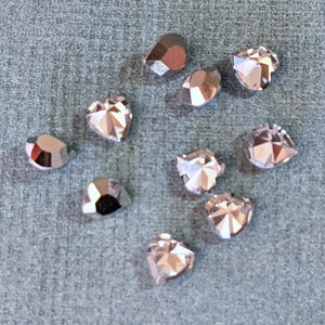May include: A collection of faceted, heart-shaped rhinestones in various sizes. The rhinestones have a metallic, iridescent finish, reflecting light with shades of silver, pink, and brown. The rhinestones are scattered on a textured, light blue surface.