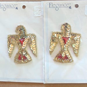 May include: Two bird-shaped patches on white backing cards labeled Elegance. Each patch features gold sequins forming the wings and body, with a central red triangular stone and red bead accents at the base. The patches are identical in design.