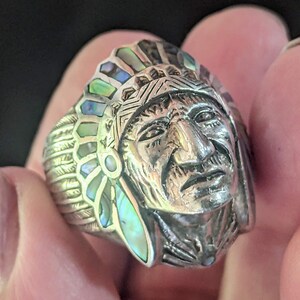 May include: A silver ring featuring a detailed Native American chief design. The ring showcases a headdress with iridescent abalone shell accents in shades of green, blue, and white. The face of the chief is intricately carved.