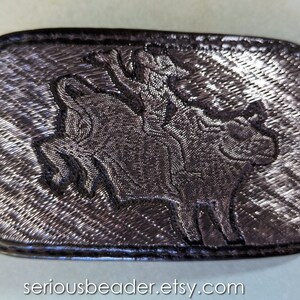 May include: A rectangular, dark gray belt buckle with a detailed embroidered design. The design features a person riding a boar. The buckle has a textured appearance and a black border. The Etsy shop name "seriousbeader.etsy.com" is visible.