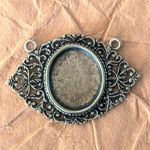 May include: An antique silver-toned oval pendant setting with ornate filigree detailing. The setting has a recessed area for a cabochon or other embellishment and two small loops for attaching a chain or cord. The background is a textured, light brown surface.