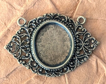 May include: An antique silver-toned oval pendant setting with ornate filigree detailing. The setting has a recessed area for a cabochon or other embellishment and two small loops for attaching a chain or cord. The background is a textured, light brown surface.