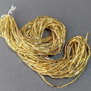 May include: A strand of gold-colored glass beads, each bead is a small cylinder. The beads are arranged in a loose coil.