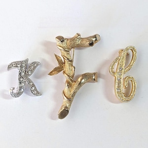 May include: Three initial letter brooches. The letter "K" is silver-toned and encrusted with rhinestones. The letter "F" is gold-toned and designed to look like a branch. The letter "C" is gold-toned and encrusted with rhinestones.