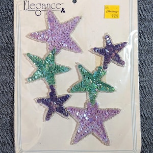 May include: A package of sequined star-shaped appliques in shades of purple and green. The stars are embellished with silver beads and are attached to a cardboard backing. The package has the word "Elegance" printed on it.