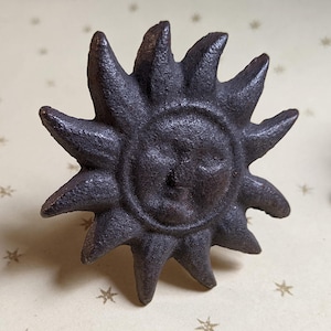 May include: A dark brown, textured metal drawer knob shaped like a sun with a central face and radiating triangular rays. The surface of the knob has a rough, cast-iron appearance, and the design features a circular center with a human-like face.