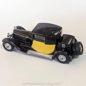 May include: A black and yellow vintage toy car with a spare tire on the back. The car has a black roof and a yellow panel on the side. The wheels are silver and the car appears to be a classic model.