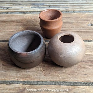May include: Three small, handcrafted ceramic pots in varying shapes and colors. One is a small, round, dark brown pot. Another is a light brown, spherical pot with a hole. The third is a small, brown, cylindrical pot. The pots are on a wooden surface.