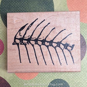 May include: A rectangular wooden stamp with a black fish skeleton design. The stamp has a light brown color and the fish skeleton is centered on the stamp. The website address "seriousbeader.etsy.com" is printed at the bottom.