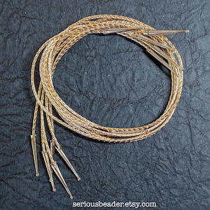May include: A coiled gold-colored braided cord necklace featuring metallic silver-toned conical aglets at the ends. The cord has a shimmering, textured appearance and is arranged in a circular loop. The text seriousbeader.etsy.com is visible.