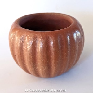 May include: A small, round, terracotta-colored ceramic bowl with a textured, ribbed exterior surface. The vessel features a wide, open top and a consistent earthy brown tone throughout, with the text seriousbeader.etsy.com printed at the bottom.