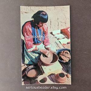 May include: Vintage photograph of a person working with clay pottery. The person is wearing a colorful top and jewelry, seated outdoors. Various pottery pieces and materials are visible, including a bowl and a small pot. The image has a warm, earthy tone.