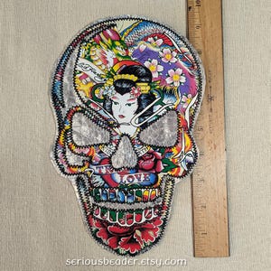 May include: A skull-shaped fabric patch featuring a colorful Ed Hardy graphic with a geisha, dragon, flowers, and the text "TRUE LOVE" and "KISS OF DEATH". A wooden ruler placed alongside shows the patch measures approximately 9 inches in height.