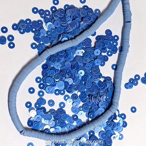 May include: A collection of small, flat, circular blue beads, including a long, continuous strand of light blue disc-shaped beads arranged in a curved line, surrounded by a scattered pile of loose, vibrant blue beads on a plain white surface.